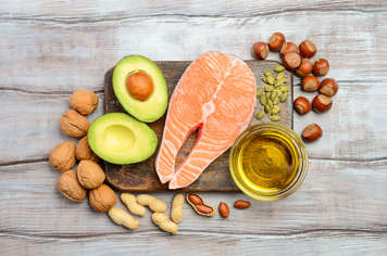 Overhead view of avocado, salmon, pumpkin seeds, and dish of oil on a cutting board with nuts arranged around the board