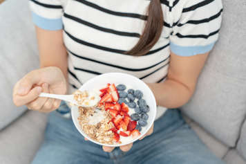 Teen girl sitting on a couch and holding a bowl of yogurt, granola, strawberries, and blueberries