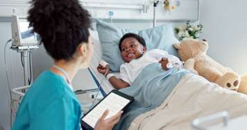 Child in a hospital bed with a pulse oximeter on his finger and an IV in his other hand. A nurse sits beside the bed
