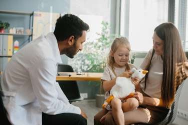 Child sitting on parent's lap across from a health-care provider who leans forward to talk to the child