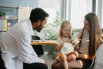 Child sitting on parent's lap across from a health-care provider who leans forward to talk to the child