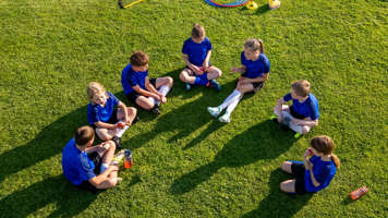 Overhead view of children in soccer uniforms sitting in a semi-circle on a grass field with sports equipment around them
