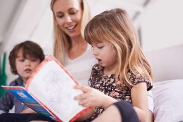 School-age girl sitting on a bed reading a book out loud. A woman and boy sit beside her